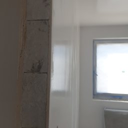 DAST - Świeżo pomalowana na biało ściana z widoczną surową kolumną z cegły, okno zabezpieczone folią malarską, fragment grzejnika i taśma malarska na ścianie.