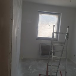 DAST - Pokój w trakcie malowania: ściany pokryte świeżą warstwą farby, zabezpieczone okno, aluminiowa drabina stojąca na podłodze z plamami farby.