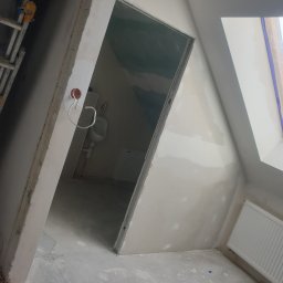 DAST - Poddasze w trakcie prac wykończeniowych: widok na surowy pokój z oknem dachowym, grzejnikiem, fragmentem drabiny i otwartym wejściem do łazienki z umywalką i toaletą, ściany po nałożeniu gładzi.