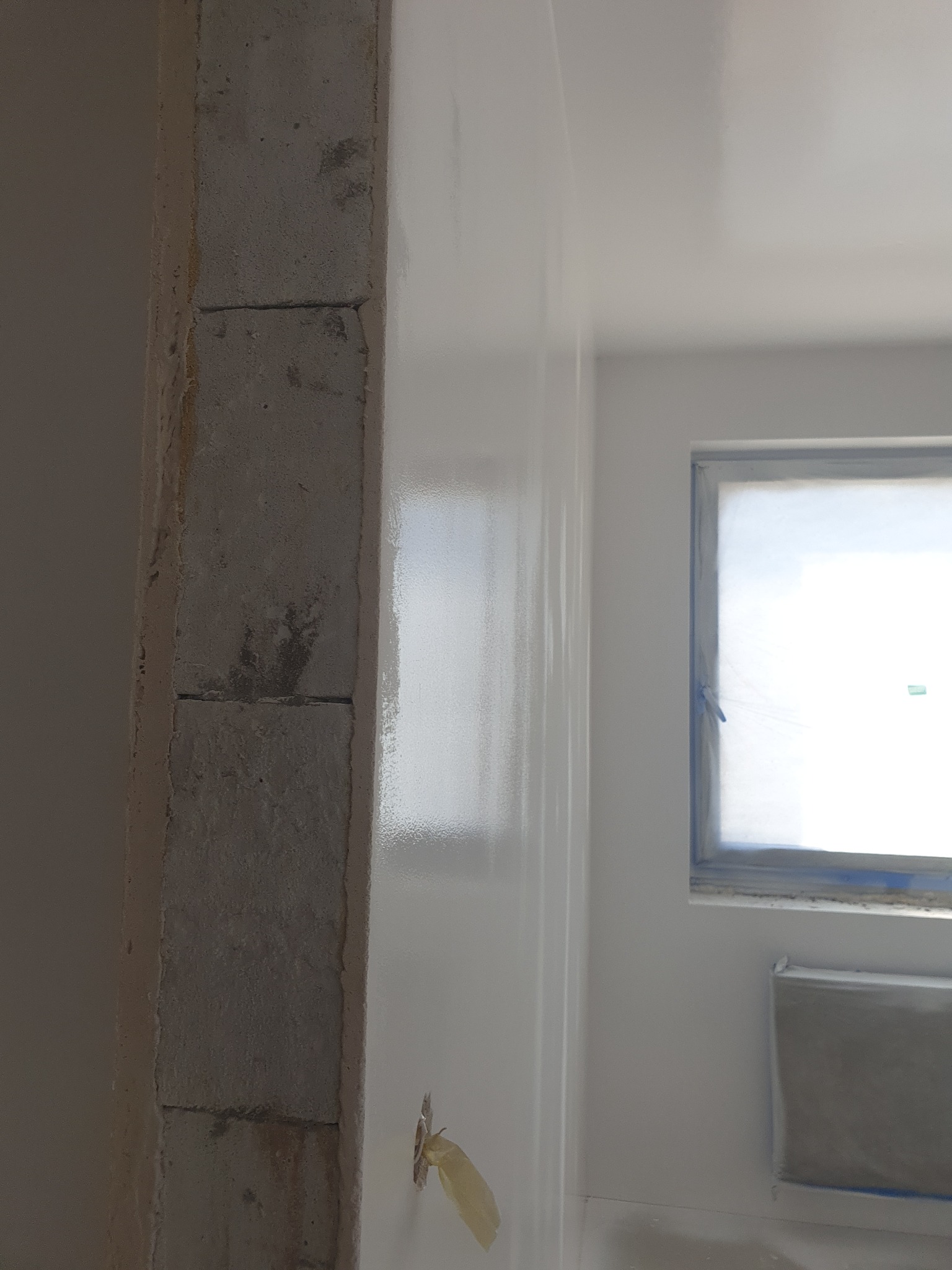 Świeżo pomalowana na biało ściana z widoczną surową kolumną z cegły, okno zabezpieczone folią malarską, fragment grzejnika i taśma malarska na ścianie.