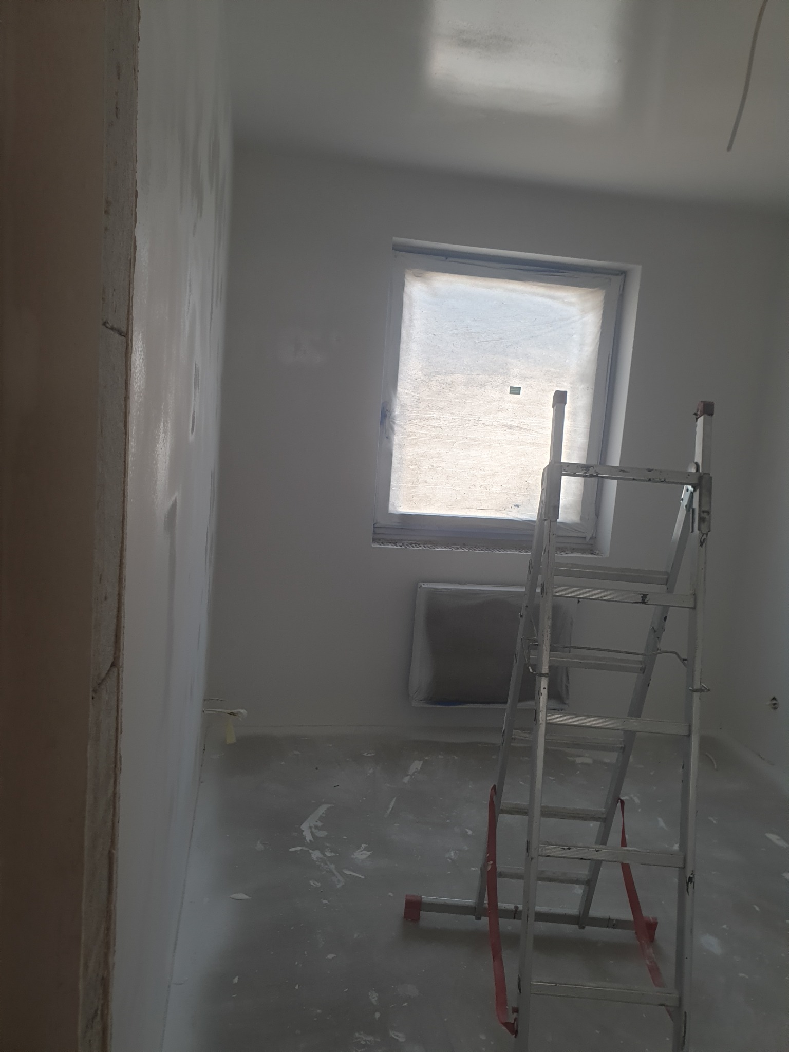 Pokój w trakcie malowania: ściany pokryte świeżą warstwą farby, zabezpieczone okno, aluminiowa drabina stojąca na podłodze z plamami farby.