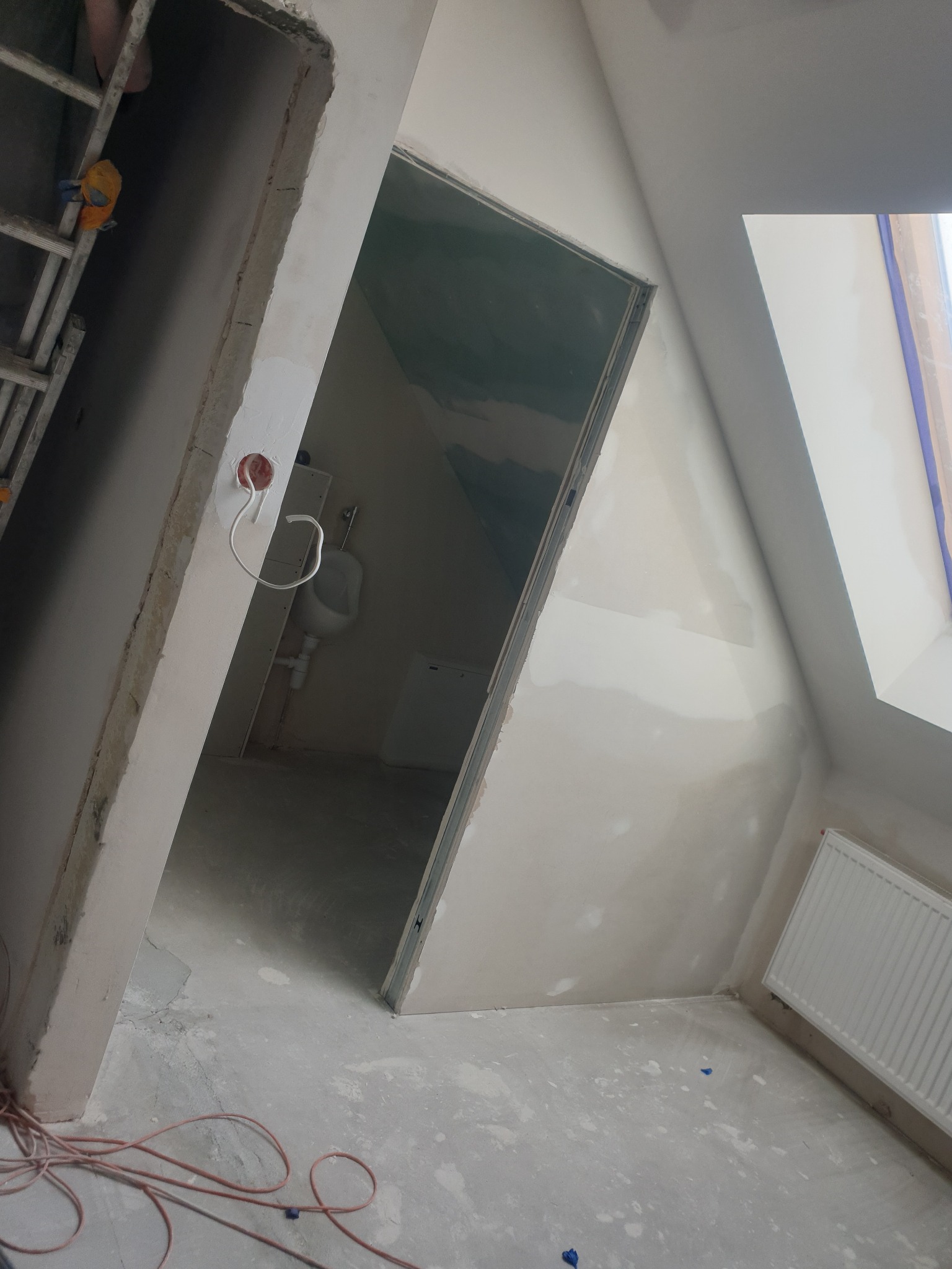 Poddasze w trakcie prac wykończeniowych: widok na surowy pokój z oknem dachowym, grzejnikiem, fragmentem drabiny i otwartym wejściem do łazienki z umywalką i toaletą, ściany po nałożeniu gładzi.