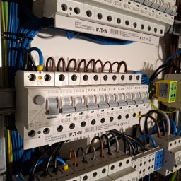 Elektron Bartosz Hodowany - Rozdzielnica elektryczna marki Eaton z widocznymi bezpiecznikami, przewodami w kolorach niebieskim, brązowym i żółto-zielonym, starannie ułożonymi i podłączonymi do listew zaciskowych.