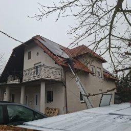 Elektron Bartosz Hodowany - Montaż paneli słonecznych na dachu dwupiętrowego domu z balkonem, pracownik na drabinie, widoczne pakiety paneli na ziemi, pochmurny dzień.