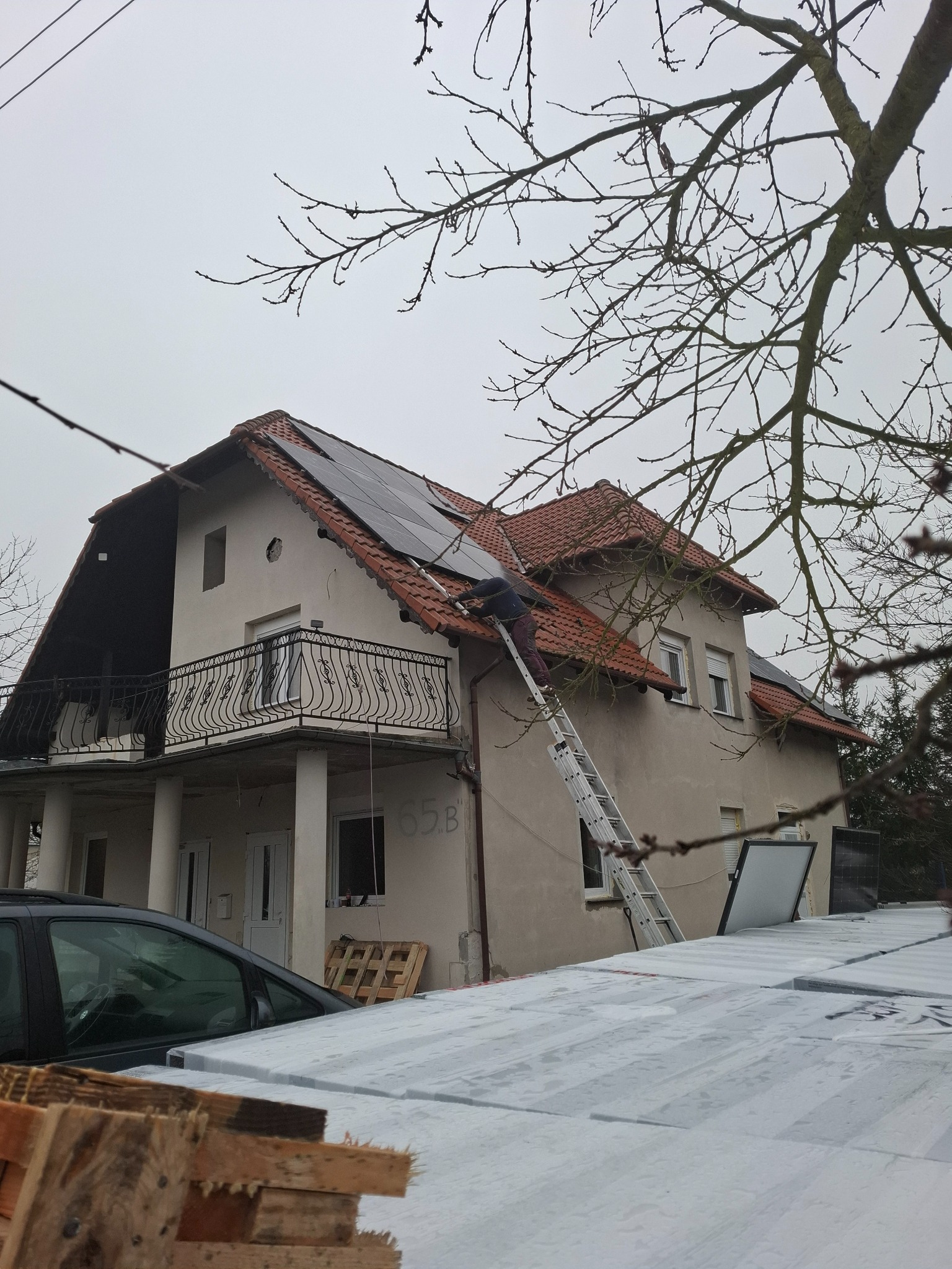 Montaż paneli słonecznych na dachu dwupiętrowego domu z balkonem, pracownik na drabinie, widoczne pakiety paneli na ziemi, pochmurny dzień.