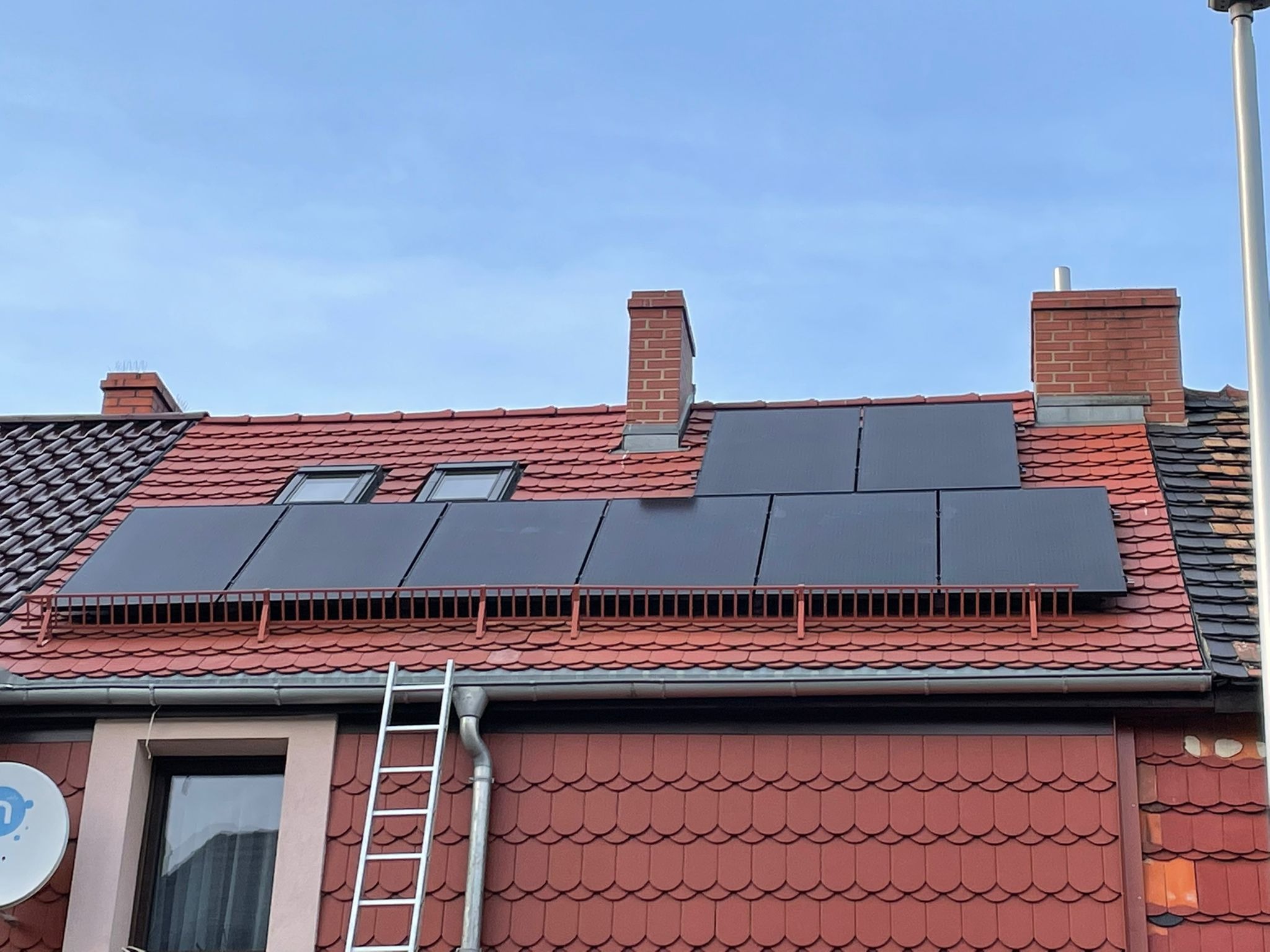 Czerwony dach domu z zamontowanymi panelami słonecznymi; drabina oparta o rynnę.