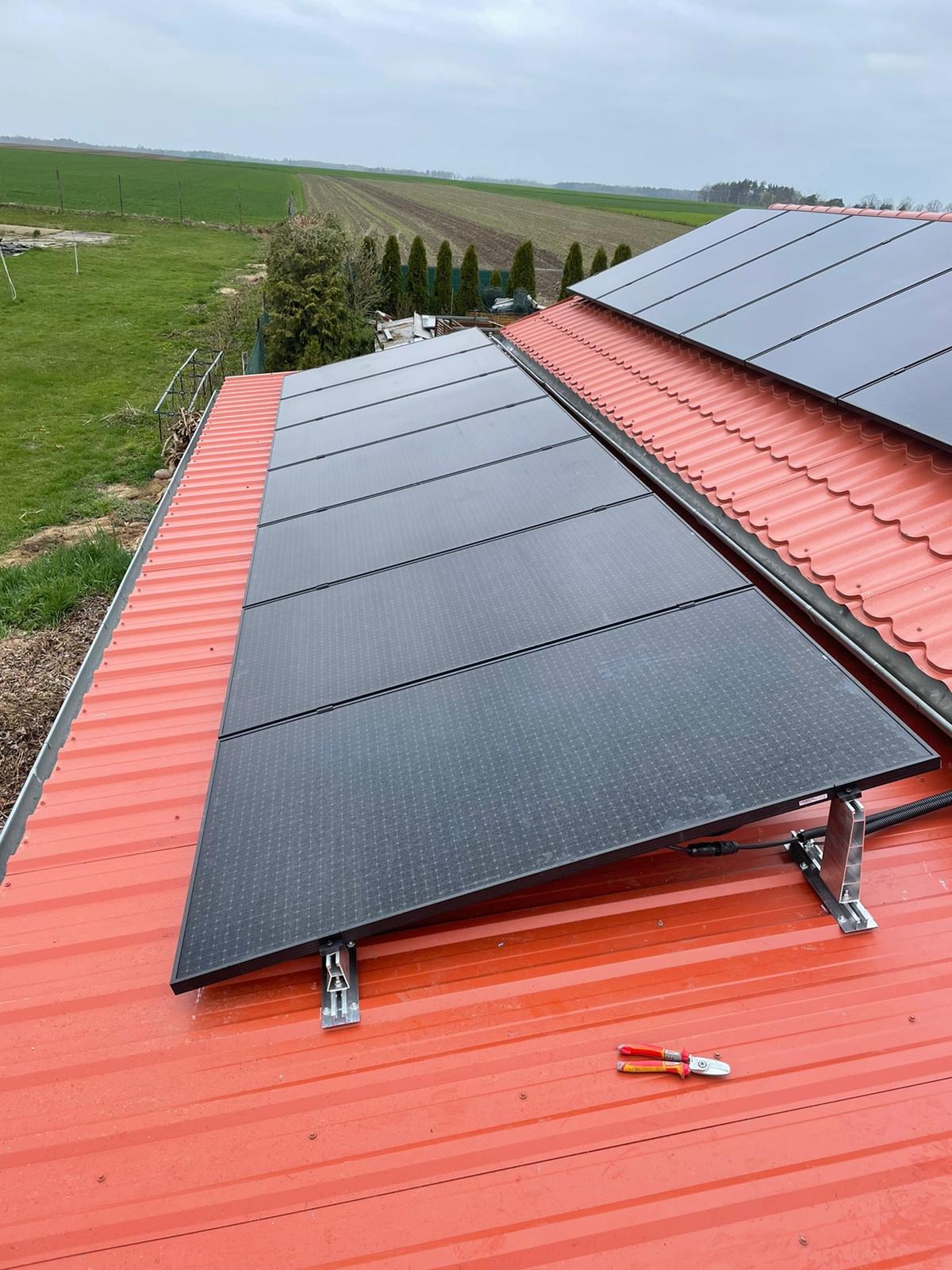 Panel słoneczny zamontowany na czerwonym blaszanym dachu, widoczne narzędzia monterskie (szczypce) oraz pole uprawne w tle.