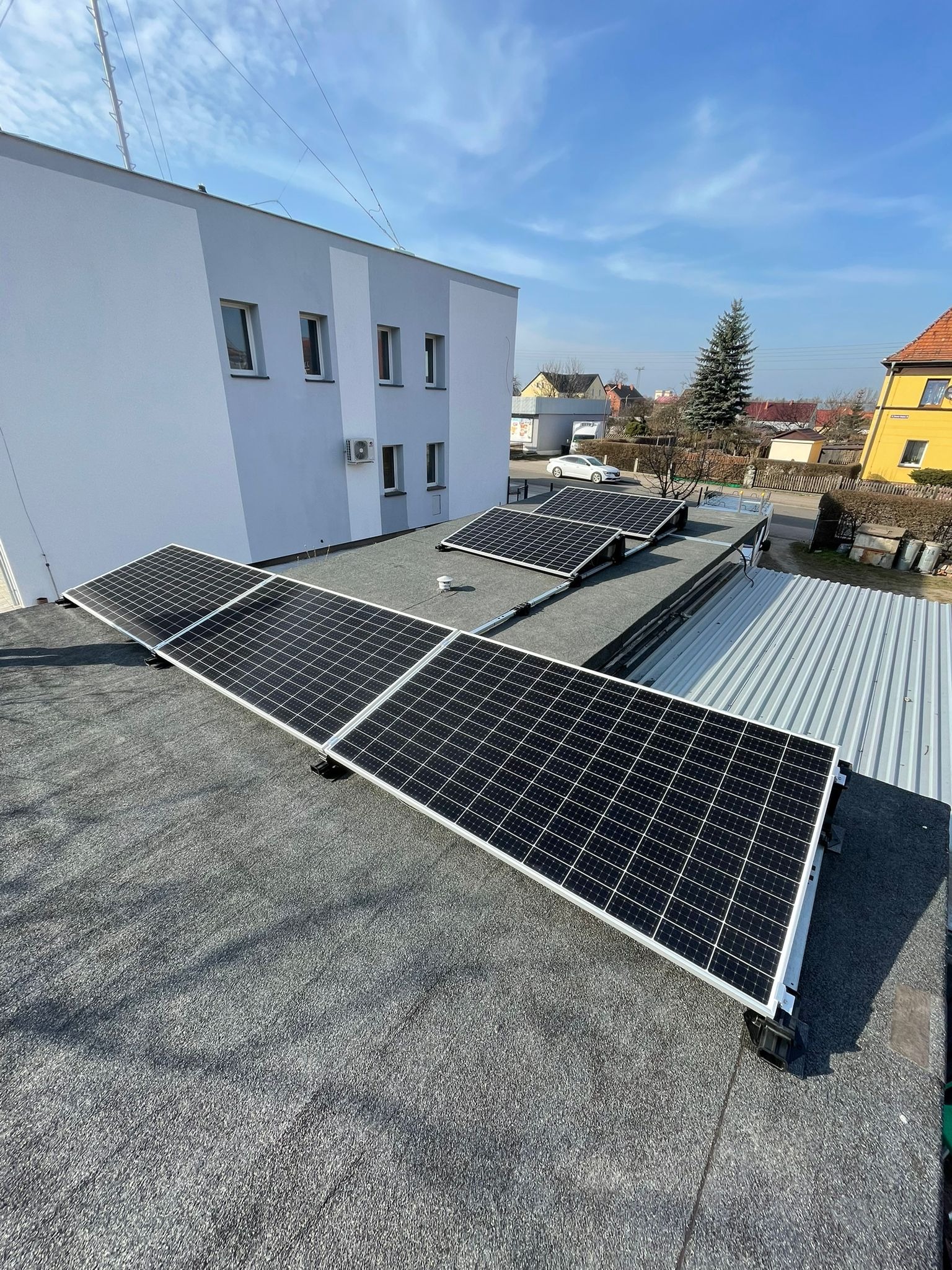 Panele fotowoltaiczne zamontowane na płaskim dachu budynku, widok z góry, w tle zabudowa miejska i błękitne niebo.