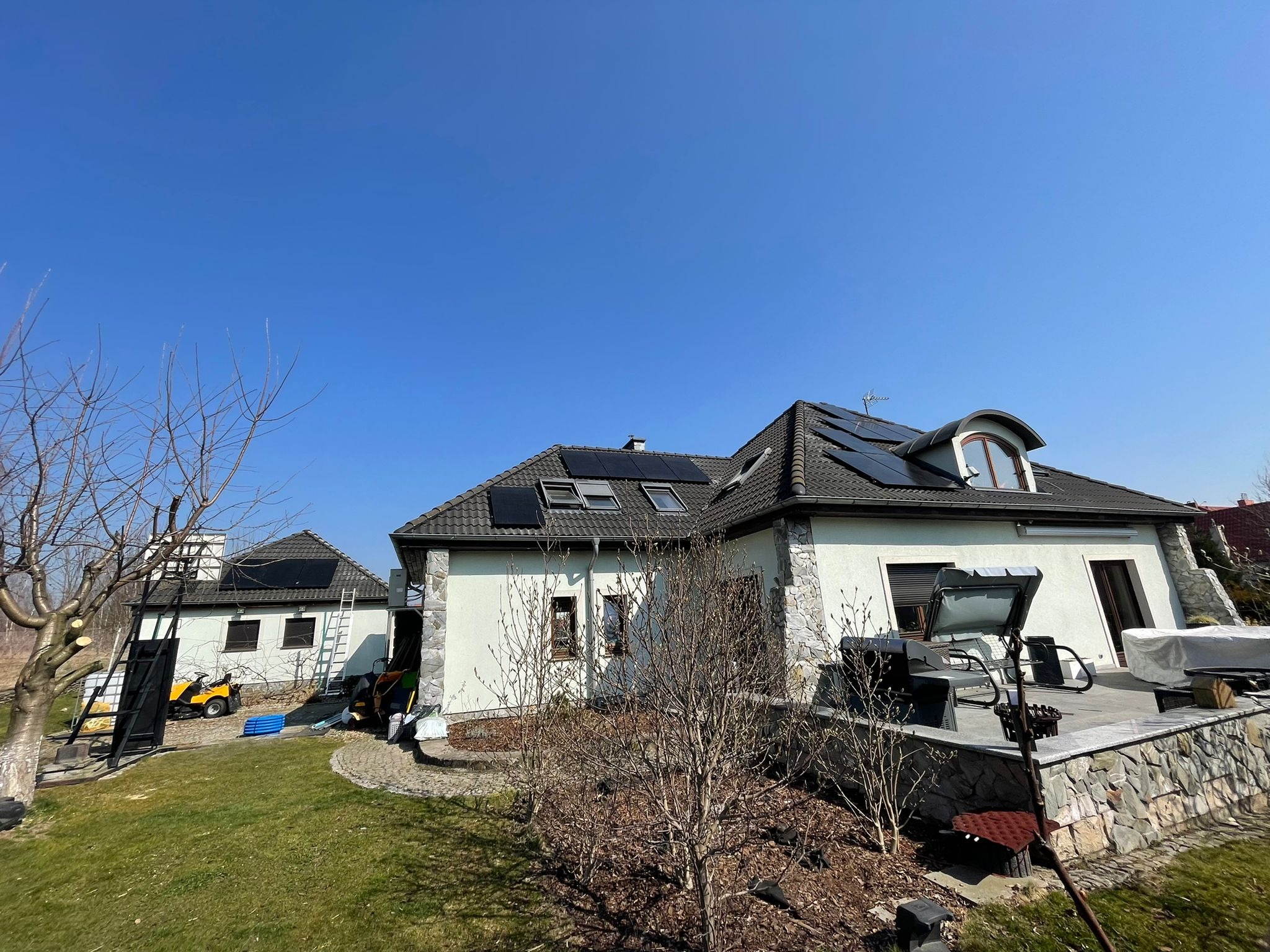 Dom jednorodzinny z panelami słonecznymi na dachu, widoczne także panele na dachu budynku gospodarczego, na trawniku sprzęt ogrodniczy i drabiny.