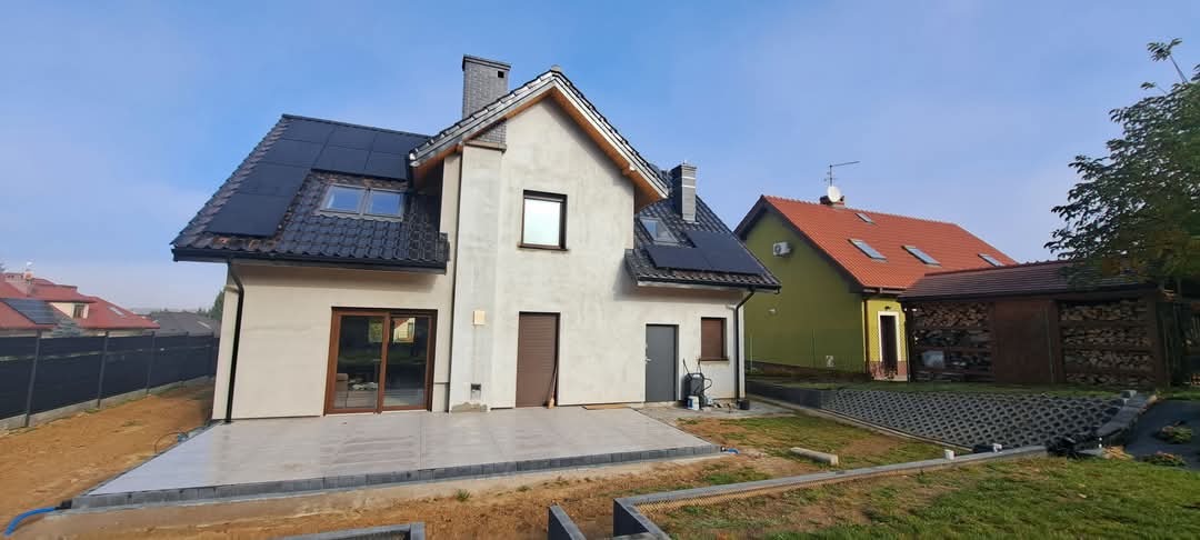 Dom jednorodzinny z panelami fotowoltaicznymi na dachu, widok z zewnątrz, w tle inne domy i drzewa, częściowo wykończony taras.