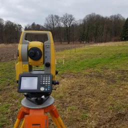 Żółty teodolit ustawiony na trójnogu na polu z młodą trawą i śladami błota, w tle widoczne drzewa i pochmurne niebo.