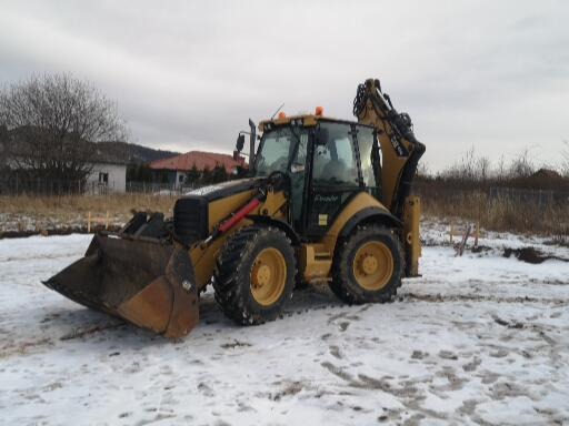 Żółta koparko-ładowarka Caterpillar stoi na zasnieżonym terenie budowy. W tle widoczny dom z czerwonym dachem i drzewa bez liści pod pochmurnym niebem. Na pierwszym planie widoczne wytyczone paliki...