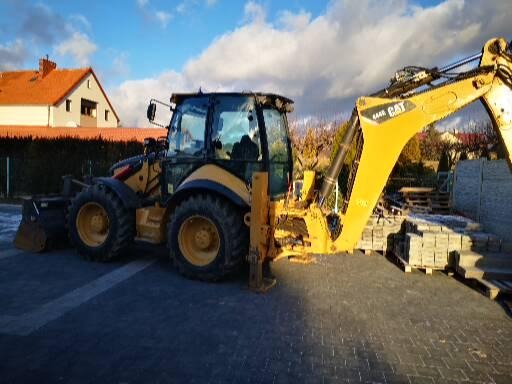 Żółta koparko-ładowarka CAT na kostce brukowej z paletami kostki w tle, w słoneczny dzień.