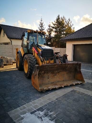 Żółta koparko-ładowarka z dużą, brudną łyżką na tle kostki brukowej, domu i garażu w słoneczny dzień.