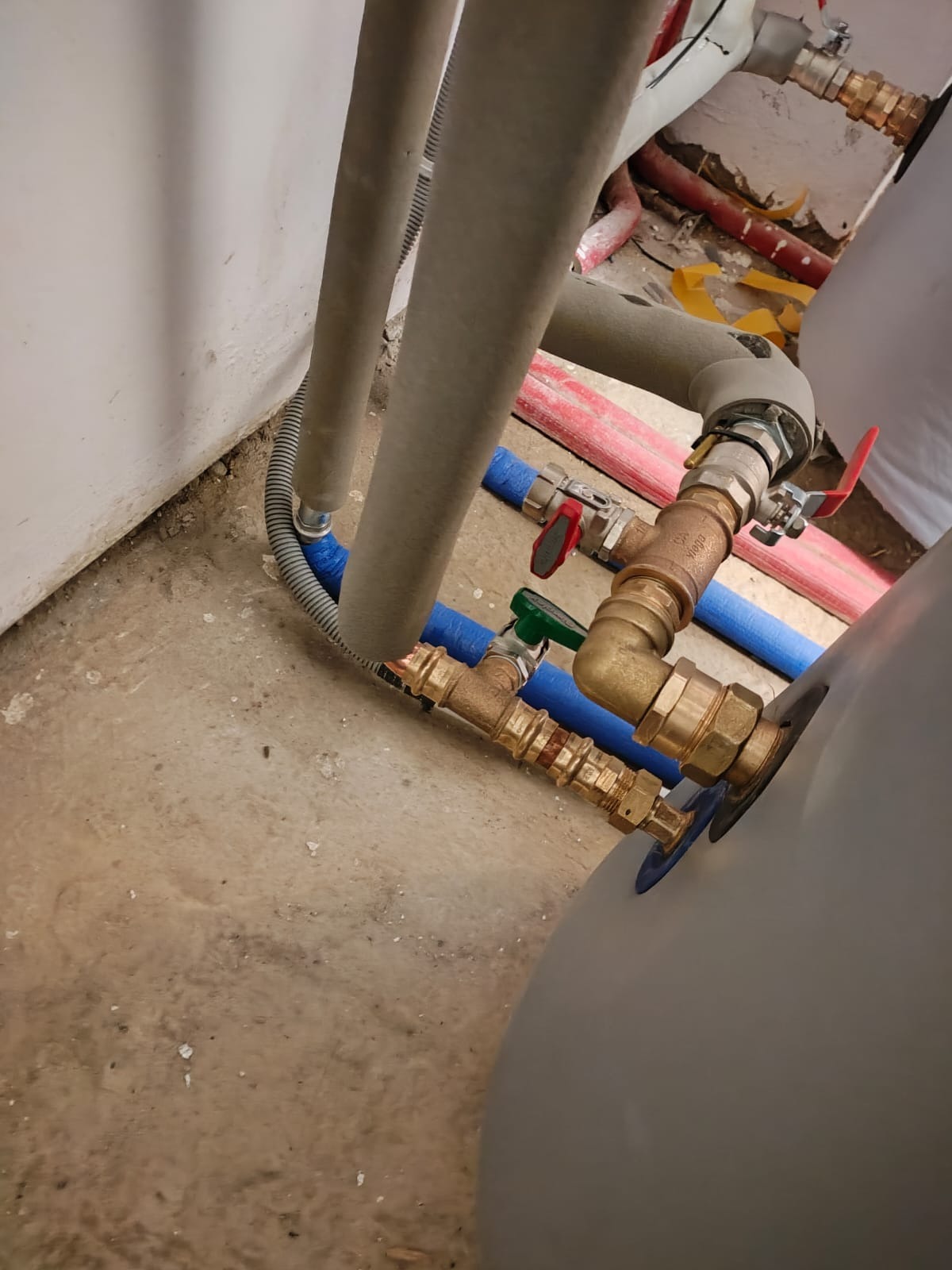 Fragment instalacji z rur miedzianych, zaworów z czerwonymi i zielonymi uchwytami, oraz izolowanych rur szarych i niebieskich, podłączonych do zbiornika w pomieszczeniu w trakcie montażu.