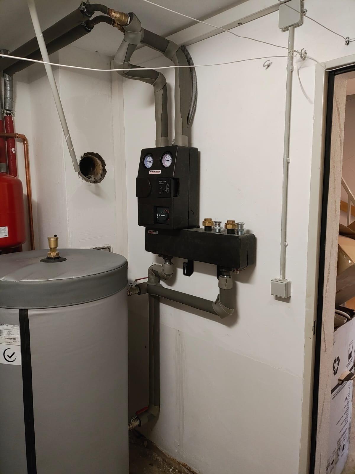 Szary zasobnik ciepłej wody użytkowej z czarnym panelem sterowania i rurami, widoczne manometry i zawory, w tle fragment czerwonego zbiornika i instalacji w pomieszczeniu technicznym.