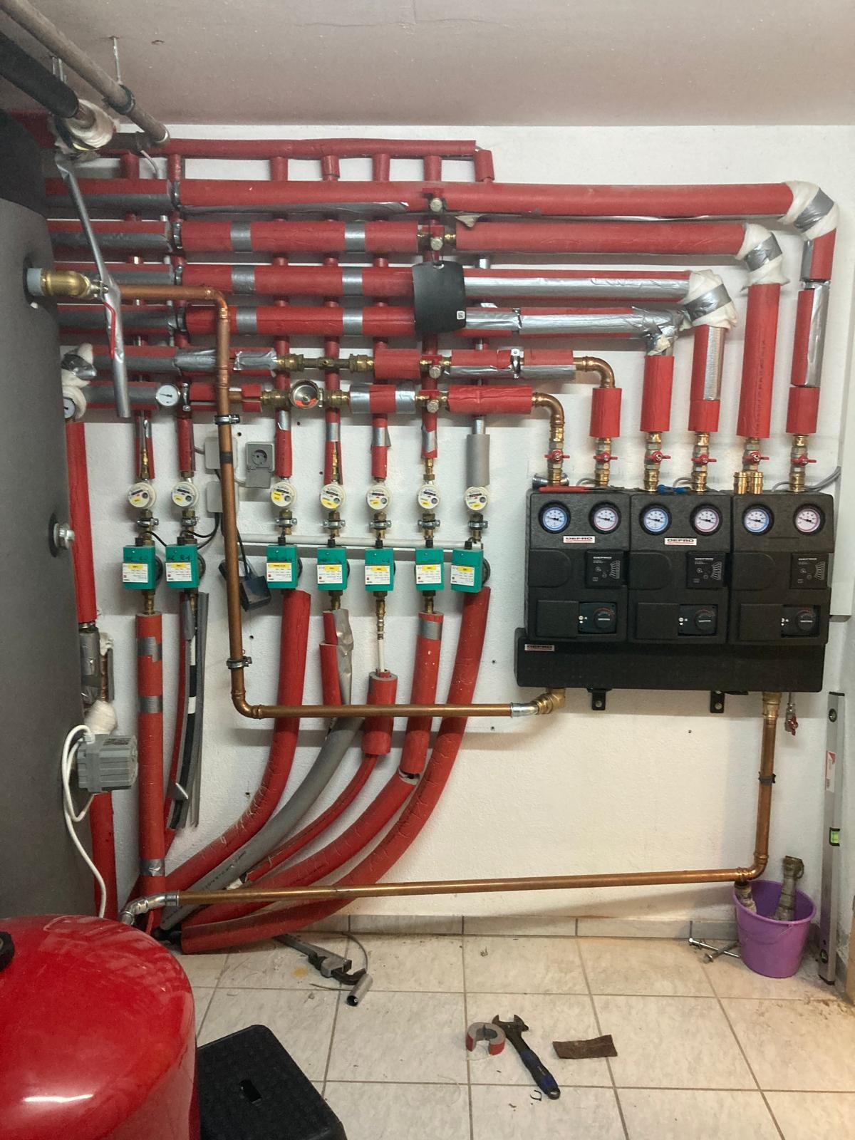 Instalacja centralnego ogrzewania z licznymi rurami, zaworami i czarną skrzynką sterującą, widoczne narzędzia na podłodze.