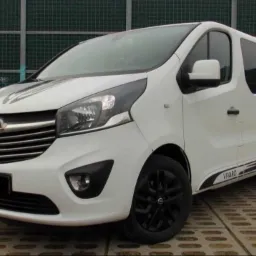 Biały Opel Vivaro z czarnymi felgami i paskami dekoracyjnymi na tle metalowego ogrodzenia z graffiti.