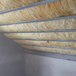 Skośny strych z widoczną izolacją termiczną z wełny mineralnej między metalowymi profilami sufitowymi, szare ściany w trakcie wykończenia.