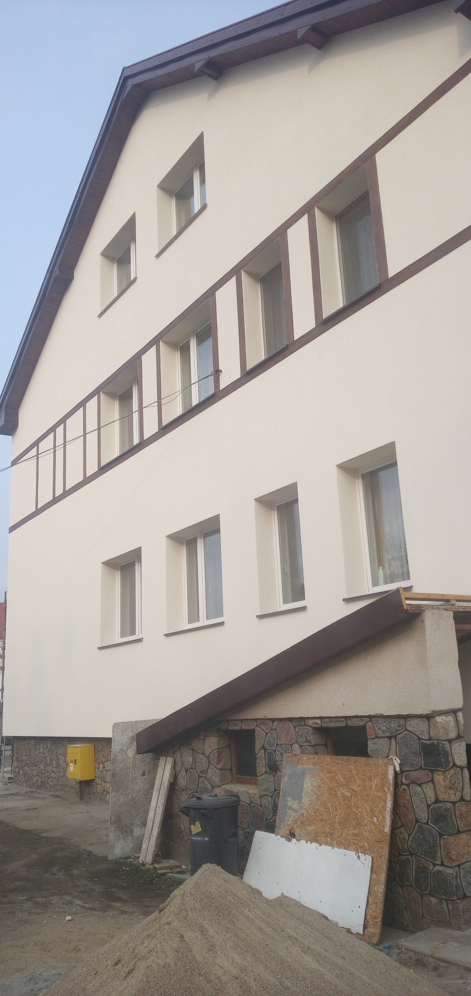 Dwupiętrowy dom z jasną elewacją i brązowymi elementami dekoracyjnymi wokół okien, widoczne materiały budowlane i śmietnik na pierwszym planie.