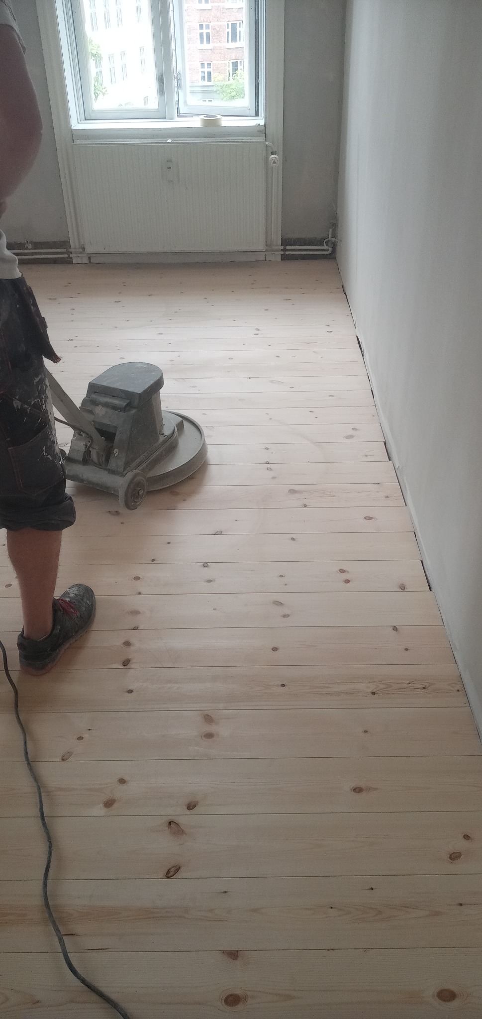 Parkiet z jasnego drewna szlifowany maszyną przez pracownika w krótkich spodniach i roboczych butach. Widoczny fragment ściany i okno z grzejnikiem.