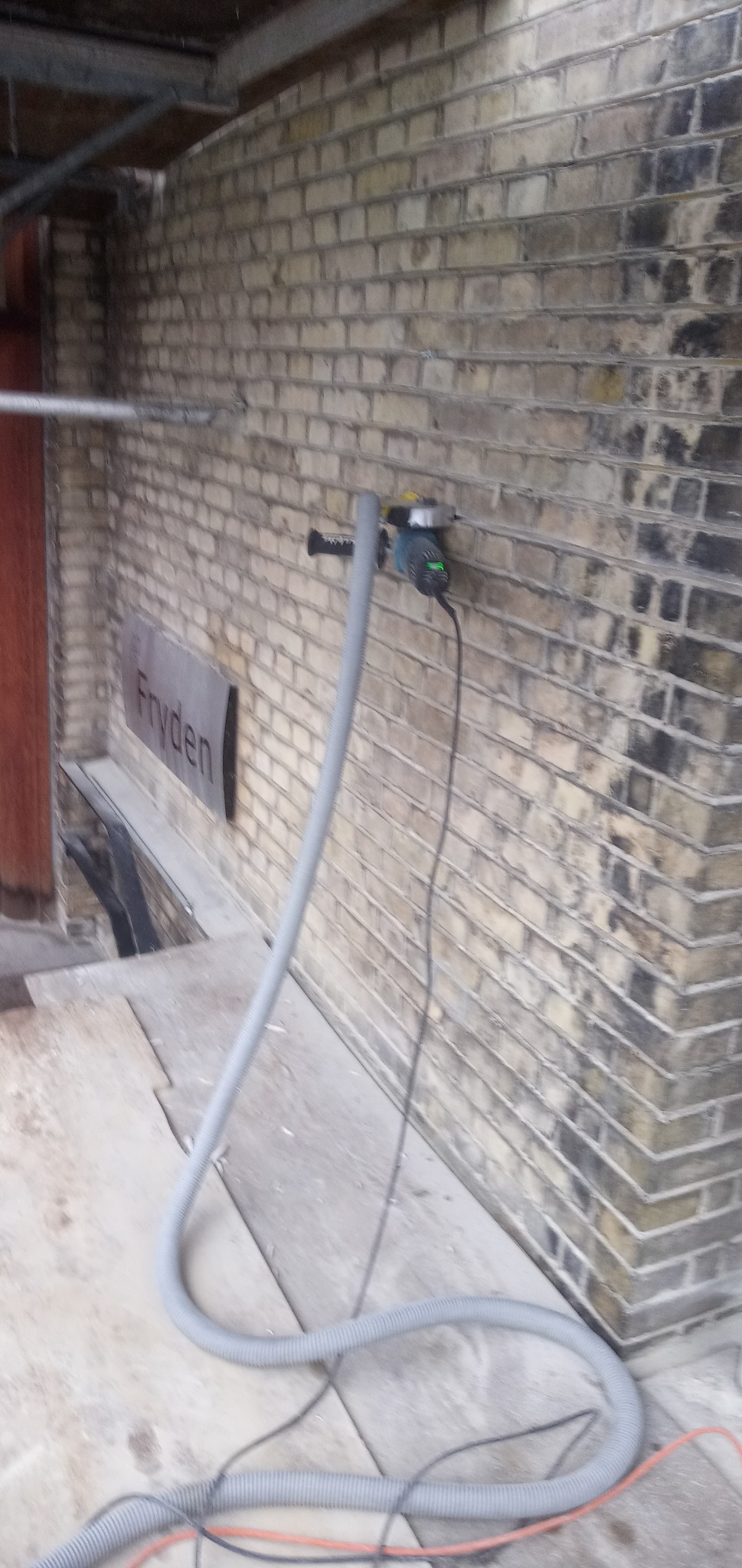 Elektryczna szlifierka z odkurzaczem przymocowana do ceglanej ściany podczas pracy, widoczny szary wąż odkurzacza i przewód zasilający.