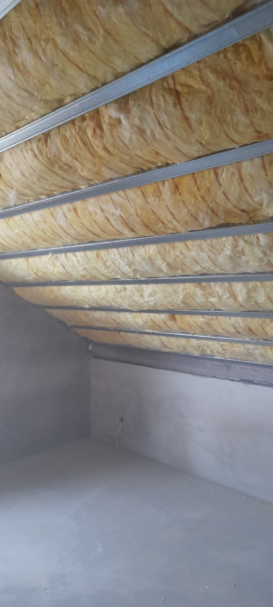 Skośny strych z widoczną izolacją termiczną z wełny mineralnej między metalowymi profilami sufitowymi, szare ściany w trakcie wykończenia.