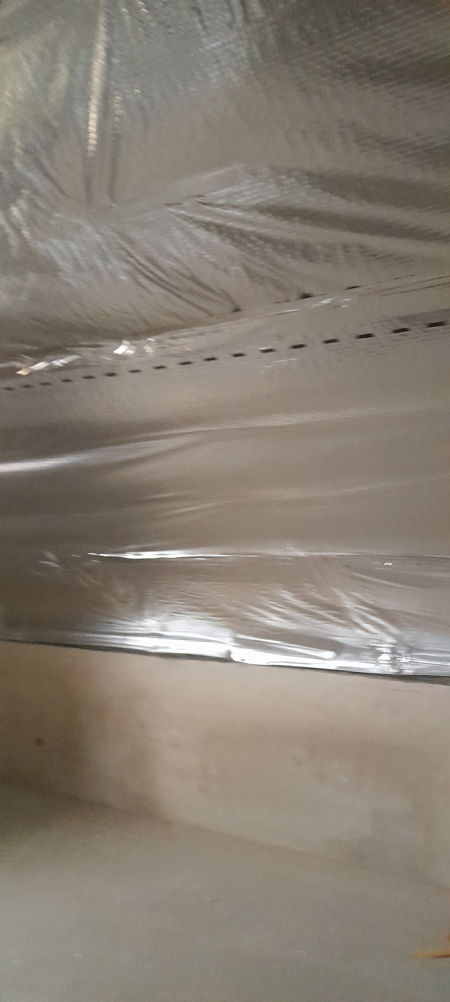 Błyszcząca, srebrna folia zabezpieczająca sufit podczas prac remontowych, z widocznymi liniami mocowania i fragmentem ściany w tle.