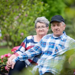 Uśmiechnięta para seniorów siedzi na ławce w parku, otoczona kwitnącymi rododendronami, mężczyzna w czapce i kraciastej koszuli obejmuje kobietę w okularach i bluzce w kratę.