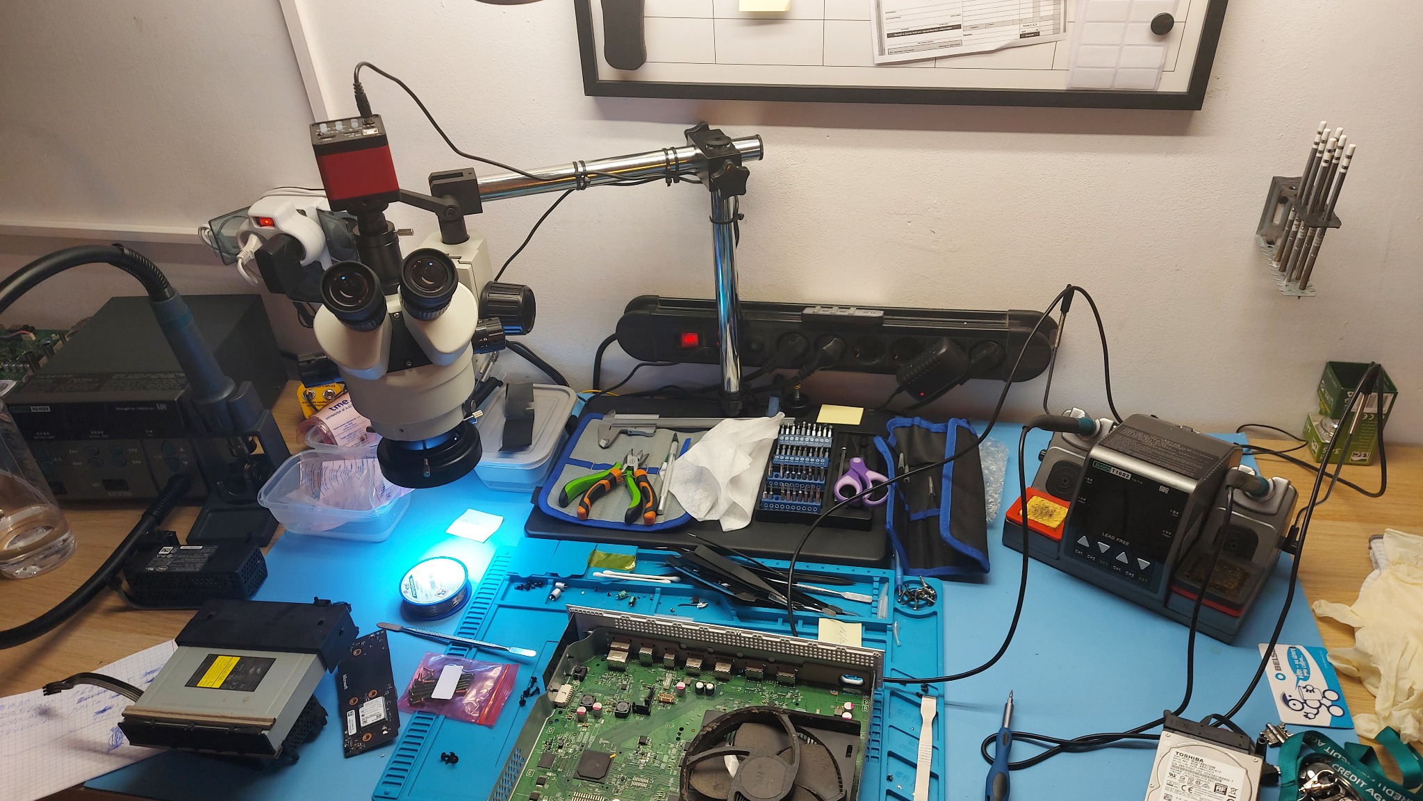 Rozłożona konsola Xbox na niebieskiej macie serwisowej, widoczne narzędzia, mikroskop, stacja lutownicza, dysk twardy i napęd DVD, sugerujące proces naprawy lub diagnostyki.