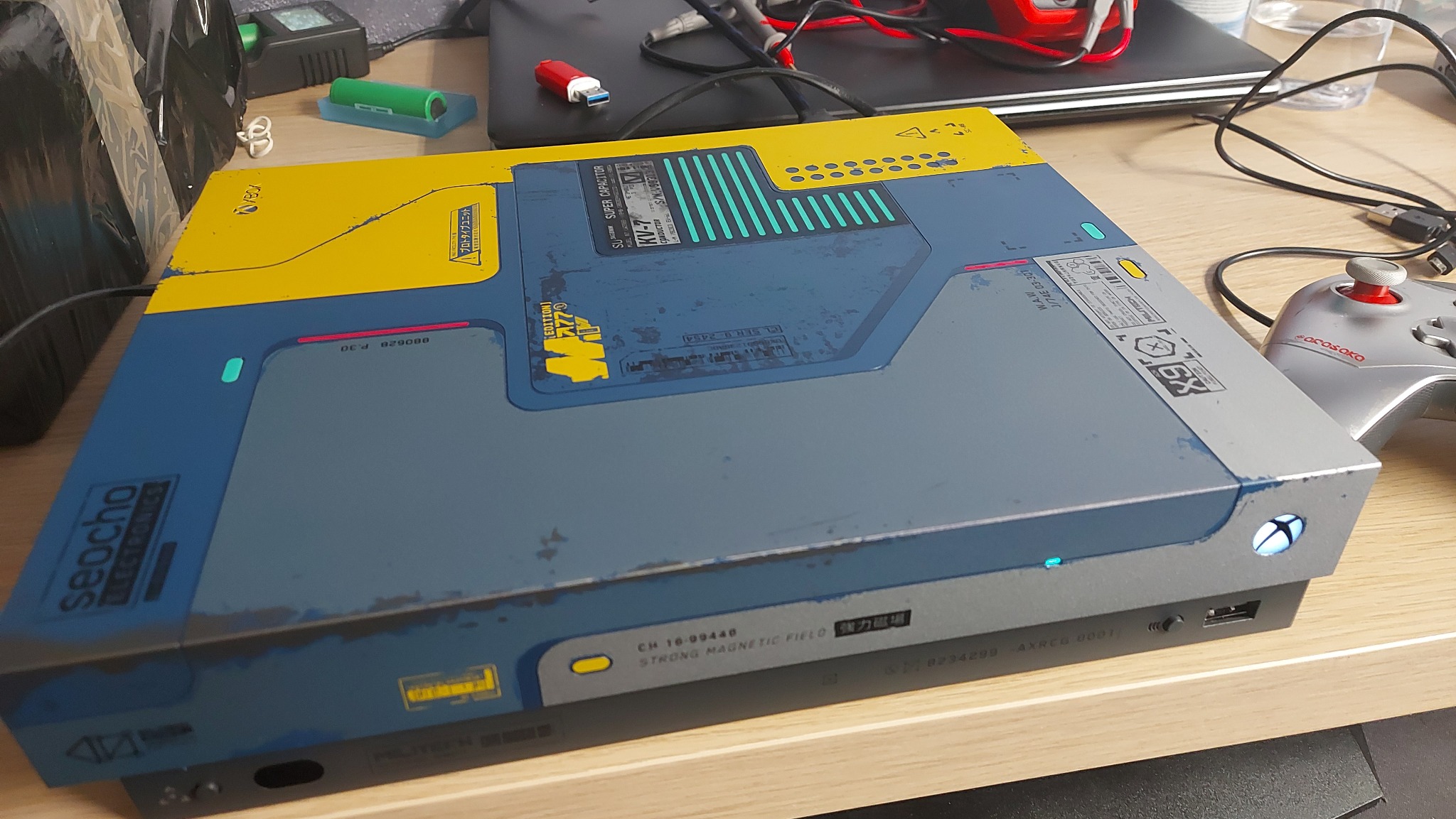 Modyfikowana konsola Xbox z motywem cyberpunkowym, z żółto-niebieską obudową ozdobioną napisami i wzorami technicznymi, leżąca na biurku obok kontrolera i innych akcesoriów komputerowych.