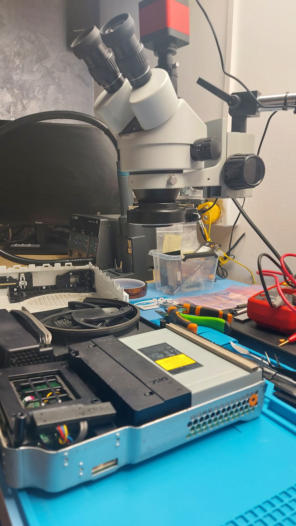 Otwarta obudowa konsoli Xbox pod mikroskopem warsztatowym, widoczne narzędzia, multimetr i elementy elektroniczne na niebieskiej macie antyelektrostatycznej.