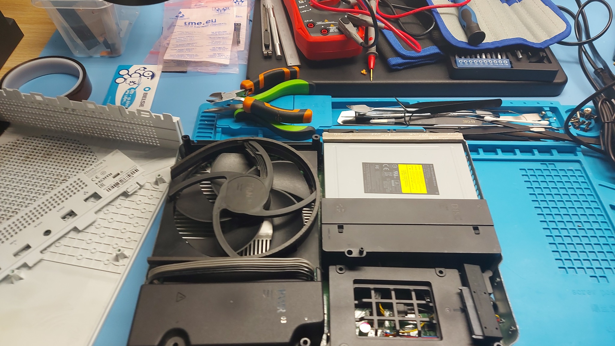 Otwarta obudowa konsoli Xbox One S z widocznym wentylatorem, napędem optycznym i częściami obudowy na niebieskiej macie serwisowej, obok narzędzi takich jak szczypce, pęsety i multimetr.