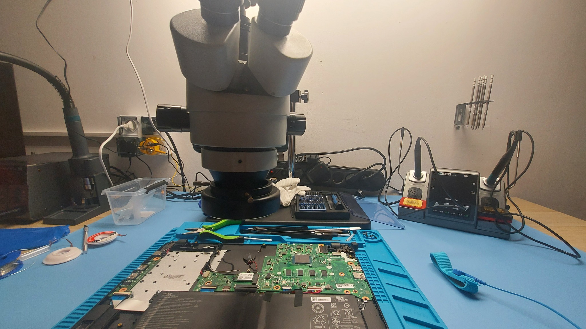Płyta główna laptopa pod mikroskopem na niebieskiej macie ESD; widoczne narzędzia, stacja lutownicza, pęsety i opaska antystatyczna.