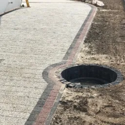Nowa nawierzchnia z kostki brukowej w odcieniach szarości i czerwieni, obrzeże z ciemnej kostki, widoczny wykop z kręgami betonowymi wypełniony wodą, świeżo przygotowany teren.