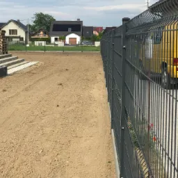 Nowo zamontowane, metalowe ogrodzenie panelowe w kolorze grafitowym, oddzielające posesję od drogi gruntowej, z widocznym fragmentem żółtego samochodu dostawczego i domami w tle.