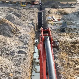 Wąski wykop wypełniony wodą, w tle czarna rura karbowana, mini koparka z przodu, widoczne ślady opon na ziemi.