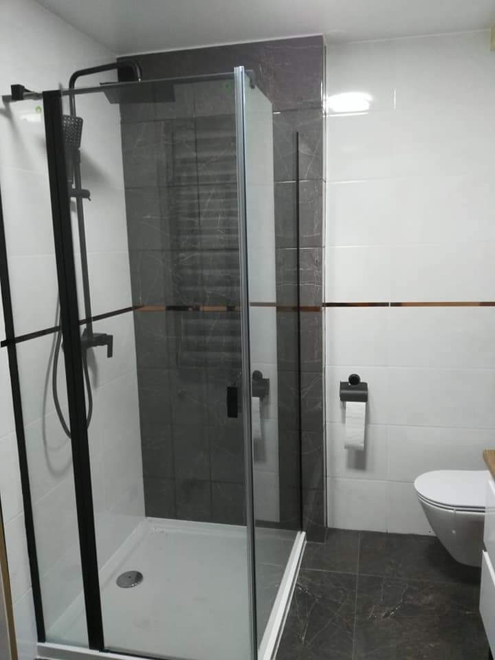 Nowoczesna kabina prysznicowa z czarnymi ramami i deszczownicą, szare marmurowe płytki na ścianie i podłodze, białe płytki z poziomym złotym paskiem, wisząca toaleta z drewnianym blatem szafki obok.