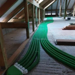 Zielone, karbowane rury wentylacyjne ułożone równolegle na poddaszu, prowadzące od rozdzielacza do otworów w dachu.