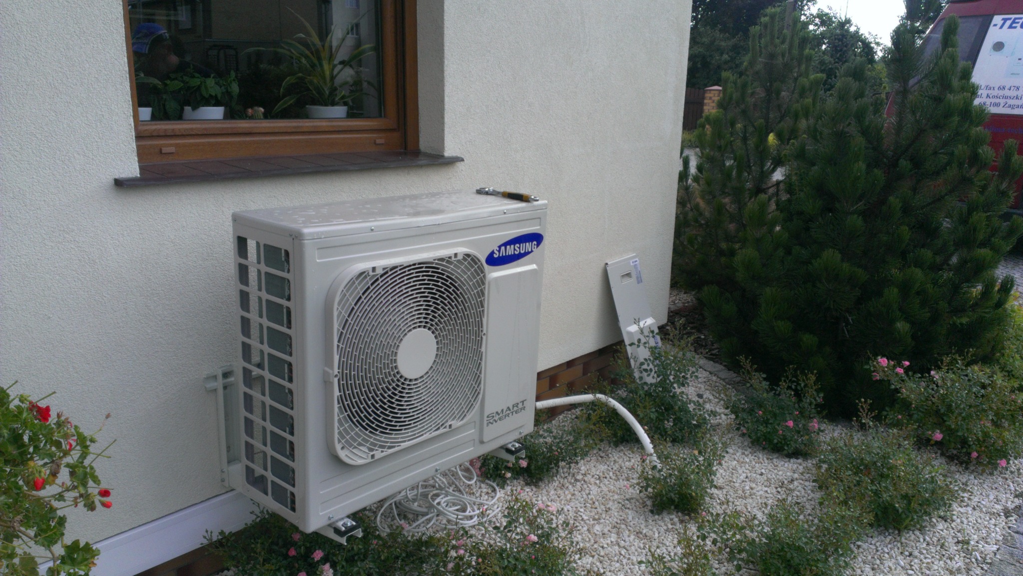 Biała jednostka zewnętrzna klimatyzacji Samsung Smart Inverter zamontowana na ścianie domu, widoczna z boku, z narzędziem leżącym na górze obudowy, otoczona kamieniami i roślinnością.