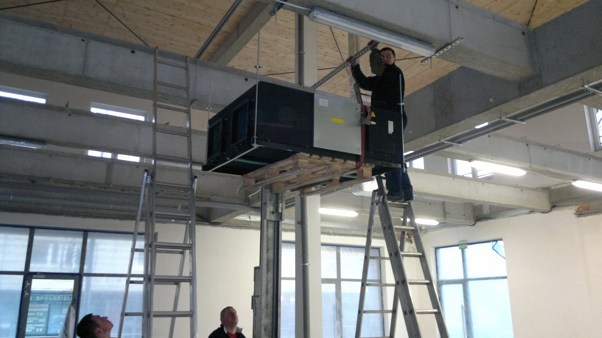 Mężczyzna na drabinie mocuje dużą centralę wentylacyjną pod sufitem w hali, widoczne elementy konstrukcyjne budynku i oświetlenie.