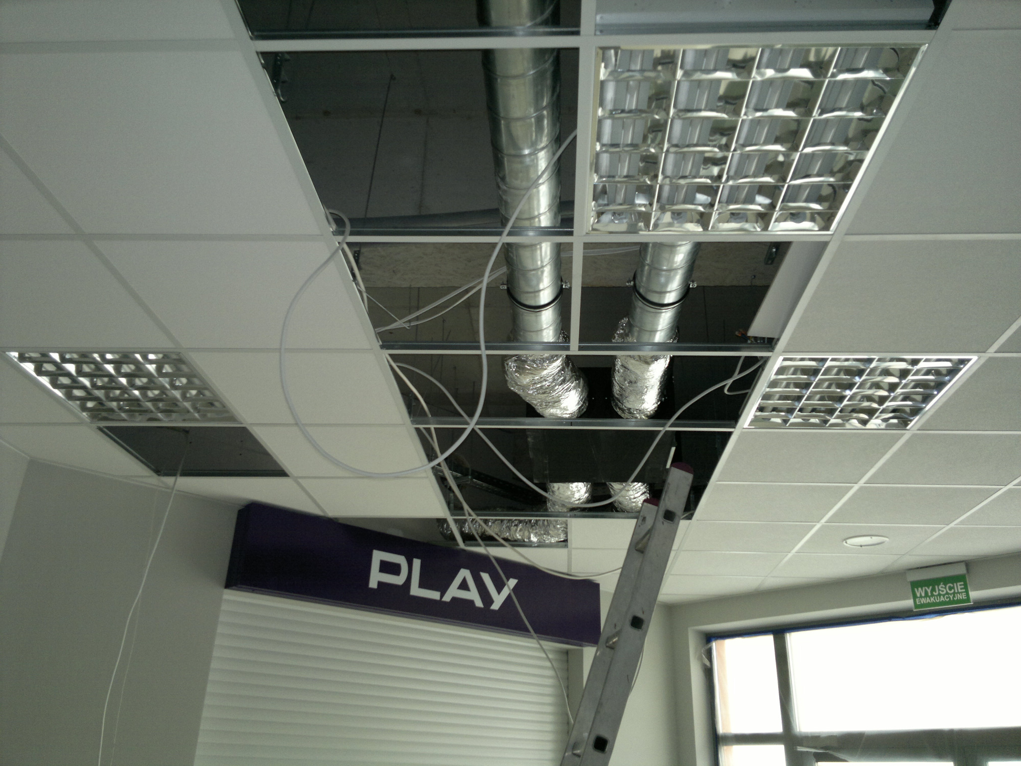 Montaż srebrnych, spiralnych przewodów wentylacyjnych w suficie podwieszanym biura z widoczną kasetonową lampą i znakiem Play oraz drabina oparta o ścianę.