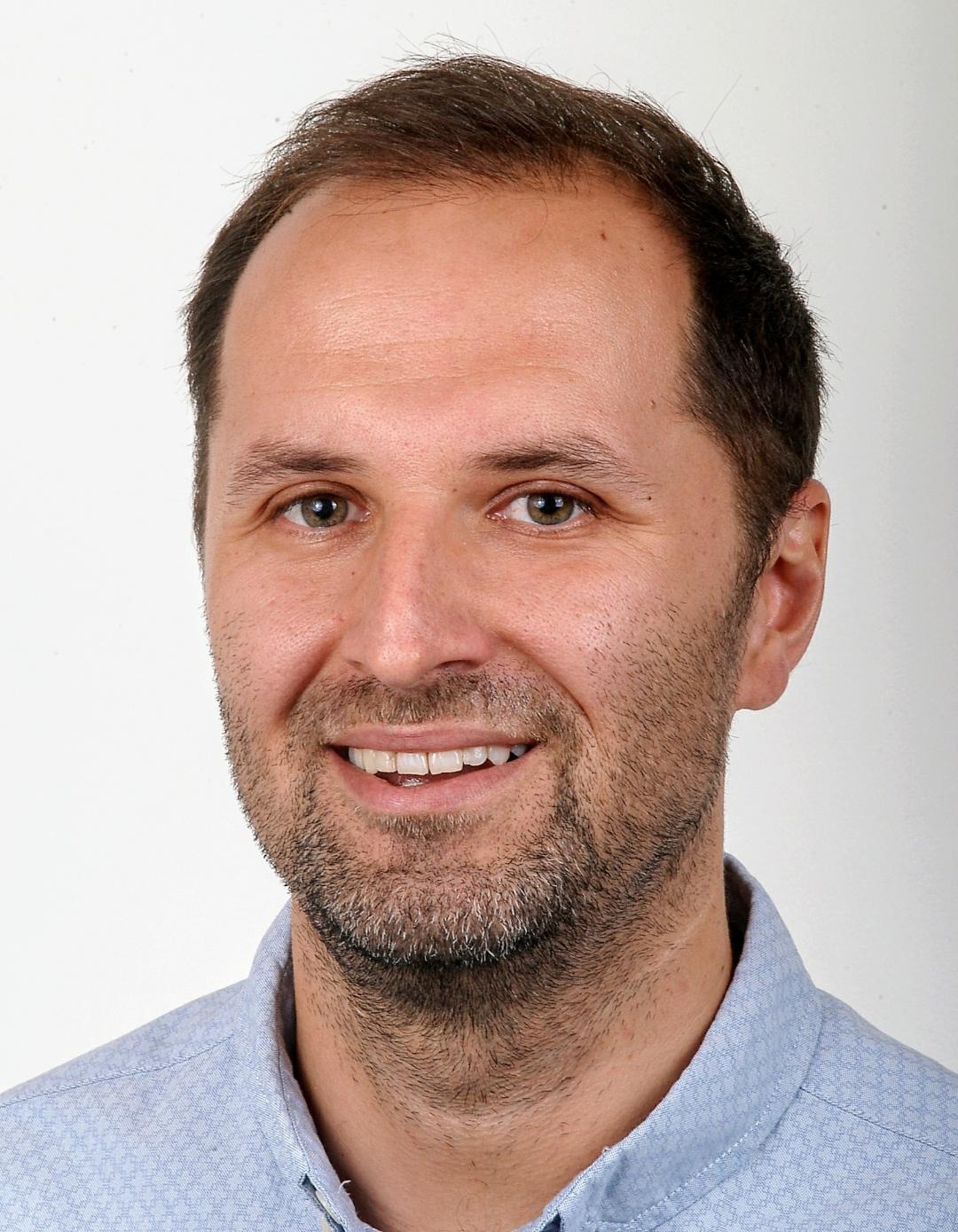 Portret uśmiechniętego mężczyzny z krótką brodą, ubranego w błękitną koszulę na jasnym tle.