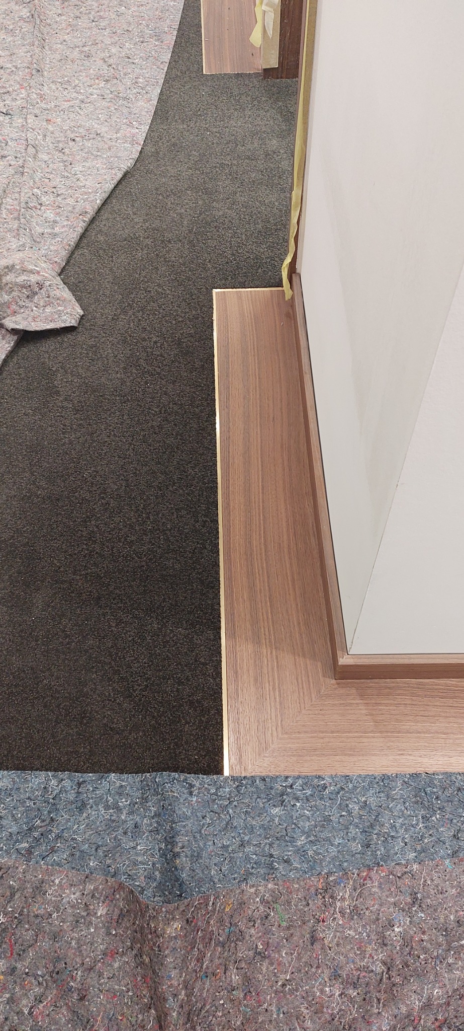 Fragment podłogi z nową, drewnopodobną listwą przypodłogową, zabezpieczoną taśmą malarską przy białej ścianie, z widocznym fragmentem szarej wykładziny dywanowej i grubego materiału ochronnego.