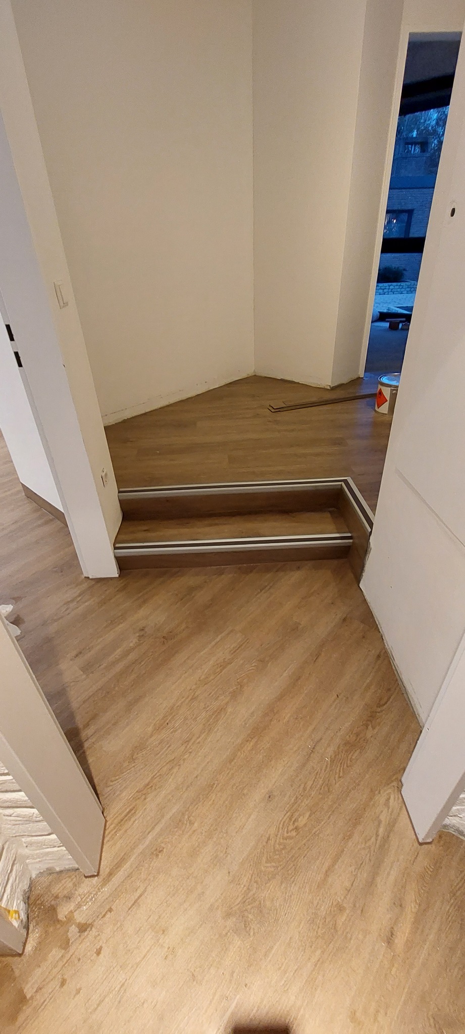 Dwa stopnie schodów wykończone panelami w odcieniach drewna z aluminiowymi listwami zabezpieczającymi krawędzie, widok z góry, w tle ściana i otwarte drzwi z widokiem na zewnątrz.