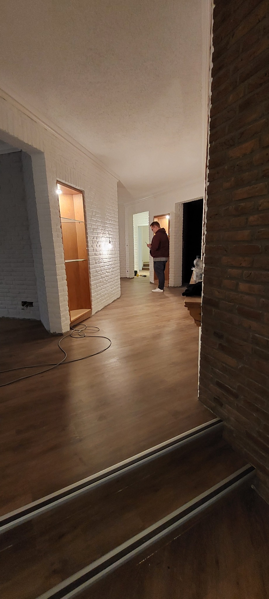 Wnętrze po remoncie z nową podłogą z paneli, widoczna osoba przeglądająca telefon, białe ściany z cegły i schody z wykończeniem.