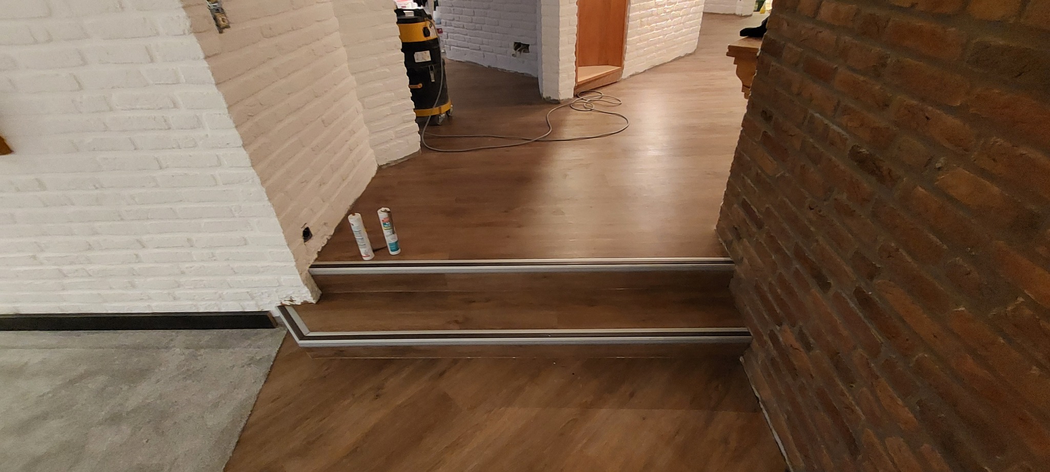 Montaż paneli podłogowych z listwami progowymi na schodach, widoczne dwie tuby kleju uszczelniającego przy ścianie z białej cegły, w tle odkurzacz przemysłowy.