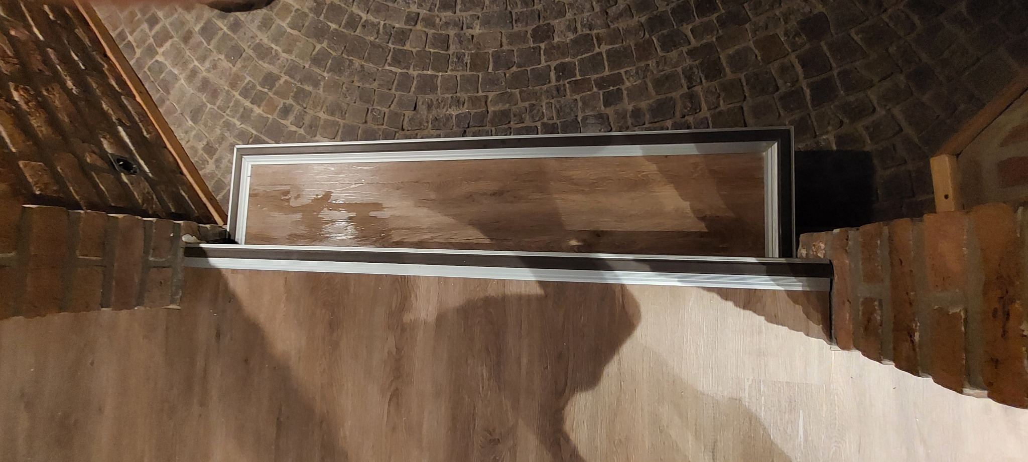 Wykończone wnętrze ze ścianami z cegły i drewnopodobną okładziną, widoczny fragment sufitu z cegły łukowej, okładzina zamontowana w ramie z metalowymi profilami.