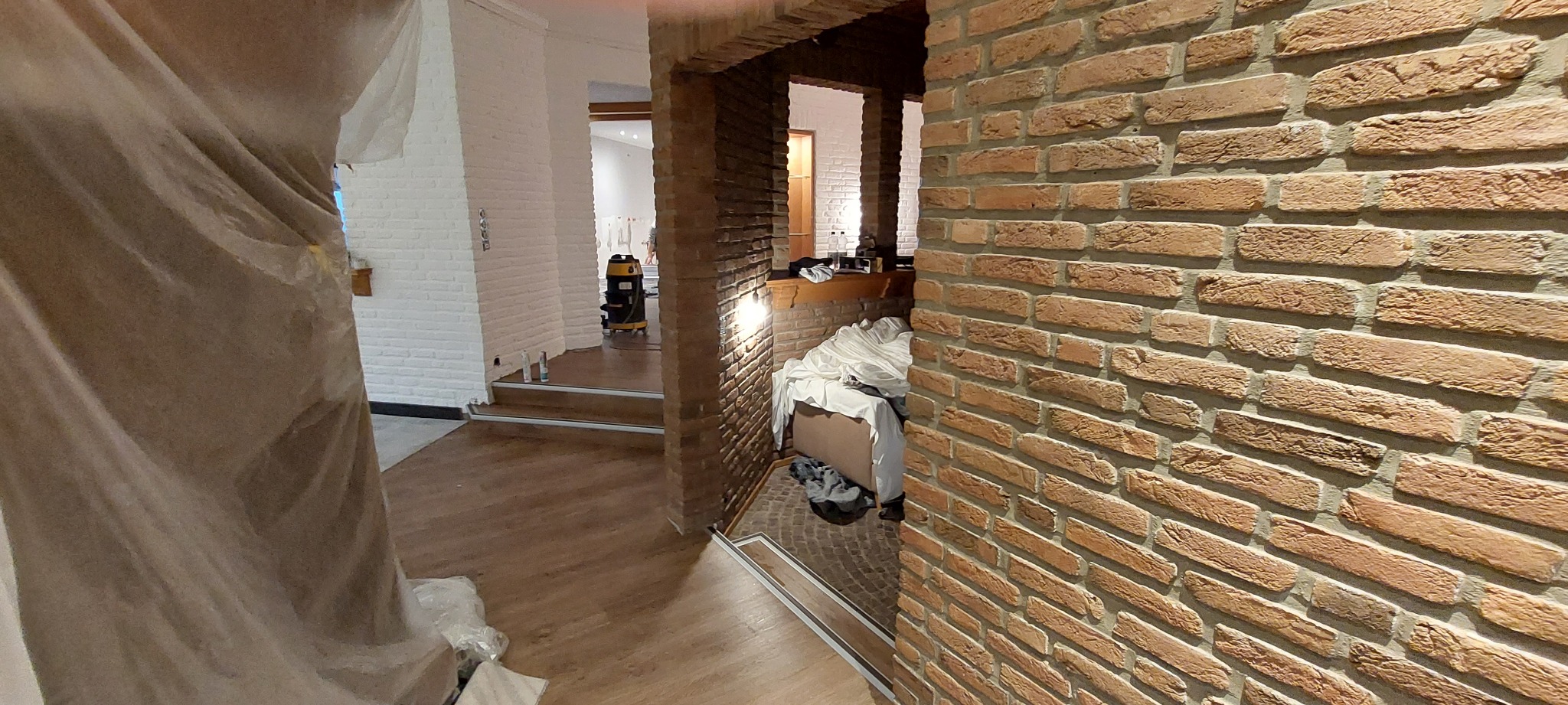 Ceglana ściana wewnątrz budynku, widok przez otwór w ścianie z widocznym pomieszczeniem w trakcie remontu, zabezpieczone folią meble oraz odkurzacz przemysłowy w tle.