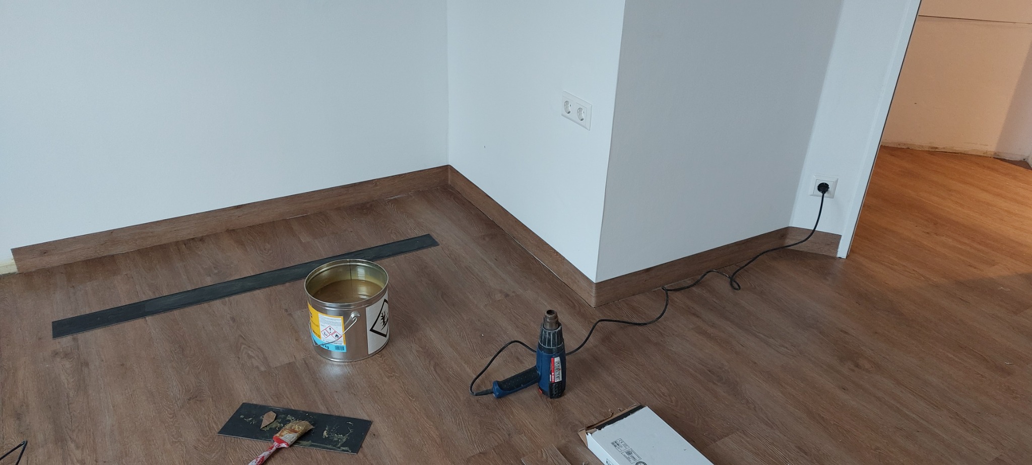 Przygotowanie do położenia paneli podłogowych: widoczna puszka z klejem, listwa przypodłogowa, opalarka, paczka paneli i narzędzia na drewnopodobnej podłodze.